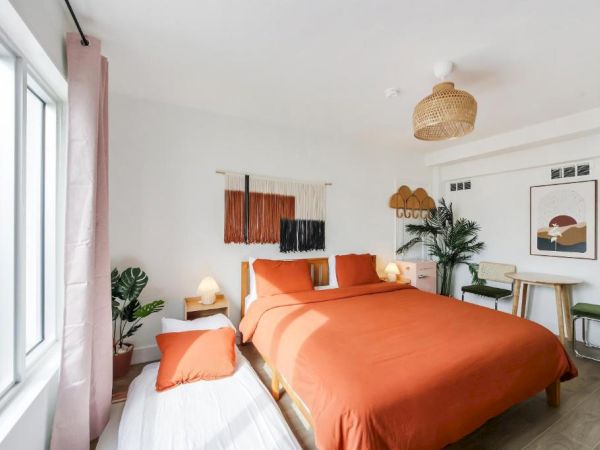 Bright modern bedroom with orange bedding, plants, and woven lights by large windows.