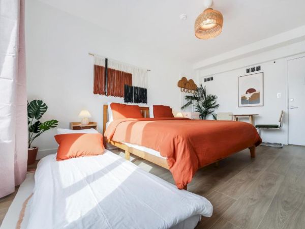 Bright modern bedroom with two beds, orange-and-white bedding, plants, wall art, and a hanging pendant lamp.