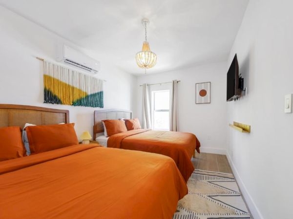 A bright bedroom with two orange beds, a hanging globe light, wall art, and a wall-mounted TV.