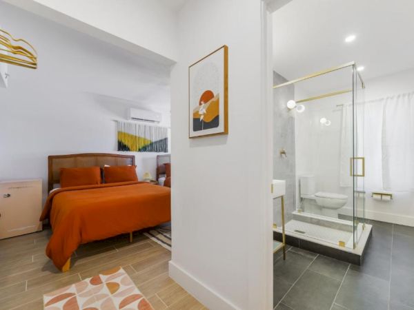 A modern bedroom with a glass-walled ensuite bathroom, featuring an orange bedspread, framed art, and a sleek toilet and vanity.