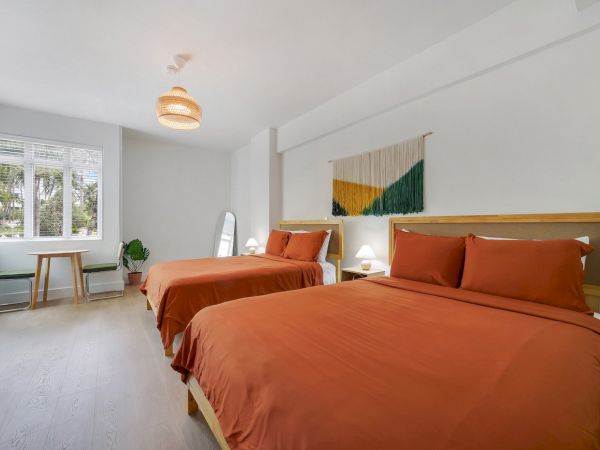 A bright modern bedroom with two beds, orange linens, a window, plants, and a colorful wall hanging.