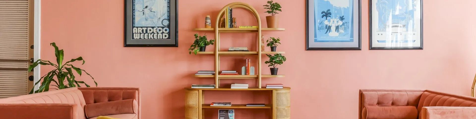 A cozy lounge with peach walls, framed art posters, potted plants, and plush orange/yellow armchairs around a small table.