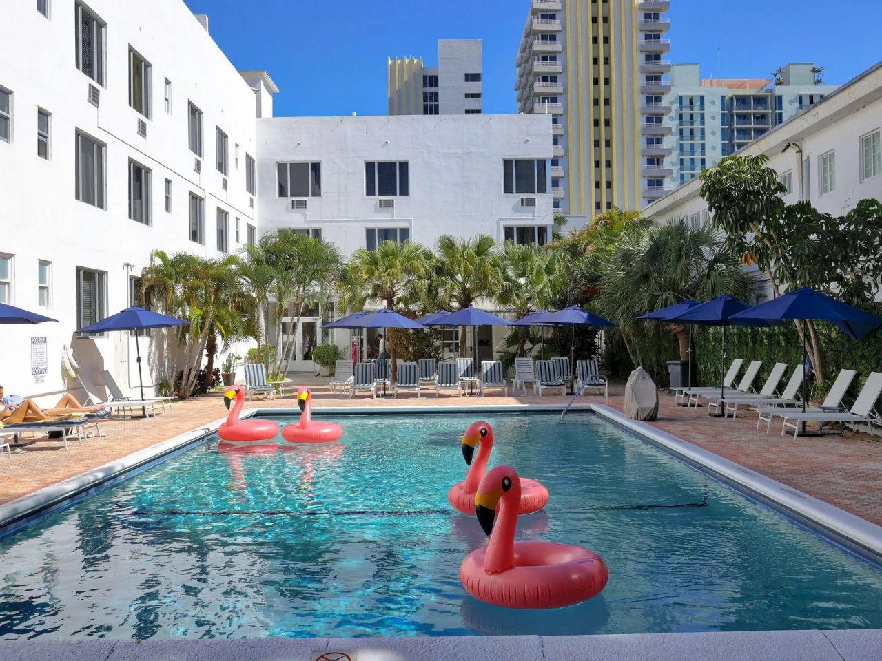 A rectangular pool with pink flamingo floats, surrounded by lounge chairs and umbrellas, in front of a white building.
