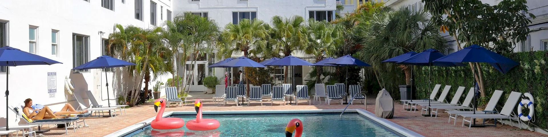 A sunny hotel pool with inflatable flamingos, blue umbrellas, lounge chairs, and palm trees in front of modern buildings.