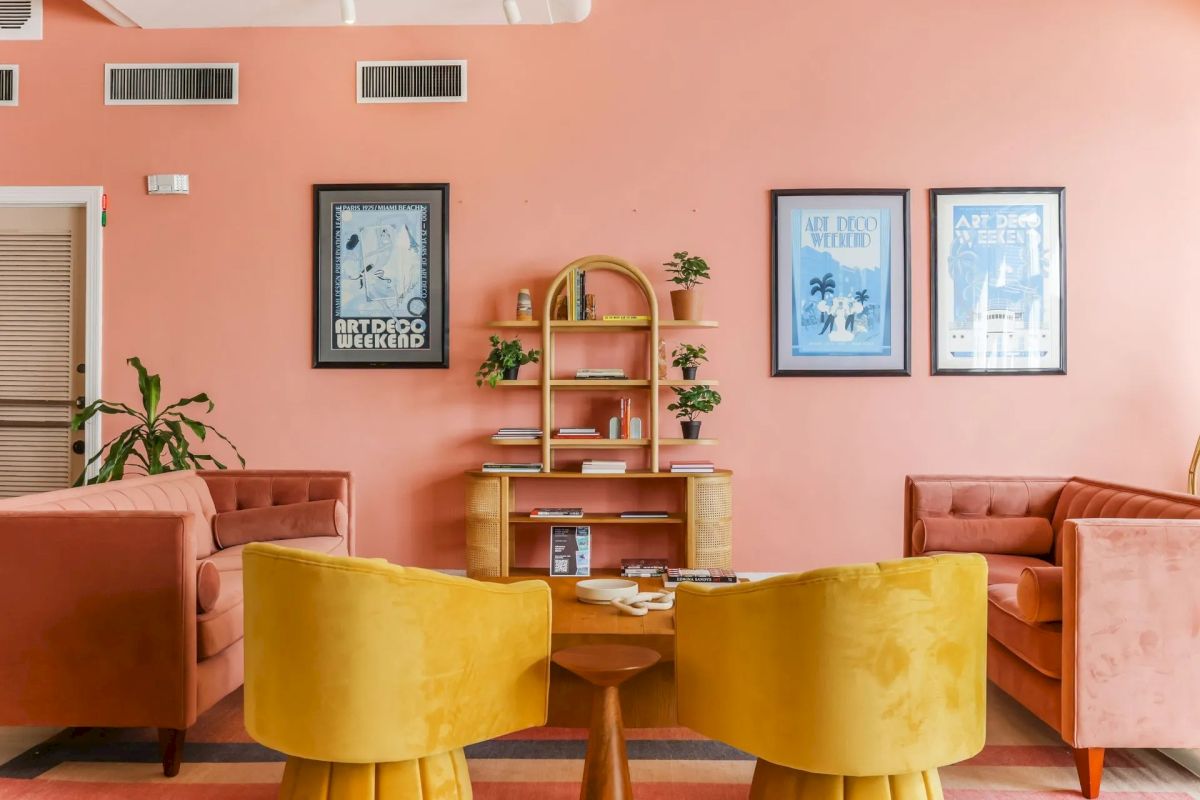 A seating area with pink sofas, yellow chairs, and a wooden table in a lobby with pink walls and framed posters.