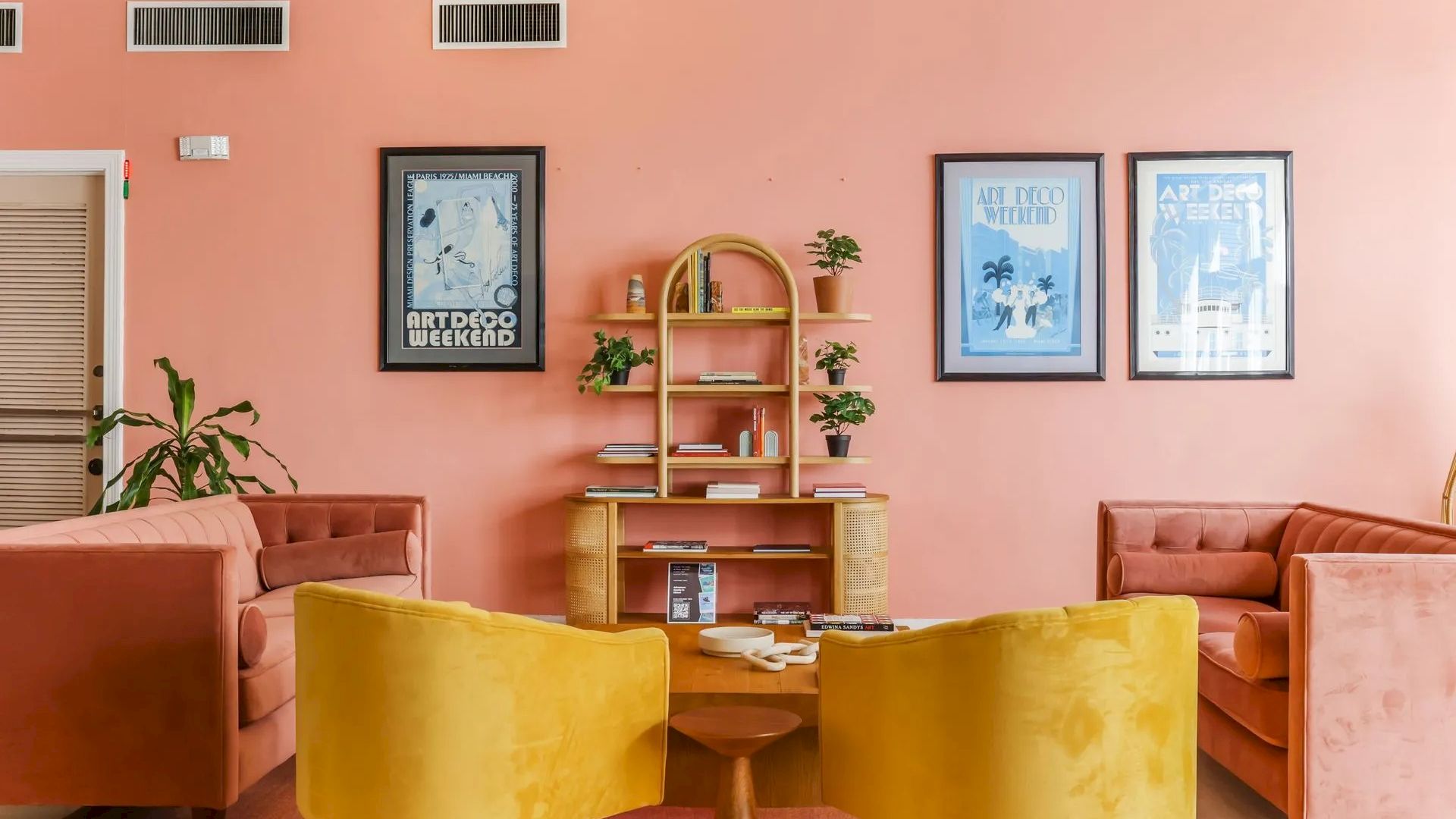 A cozy lounge area with velvet sofas, yellow chairs, a wooden table, and framed posters on a pink wall.