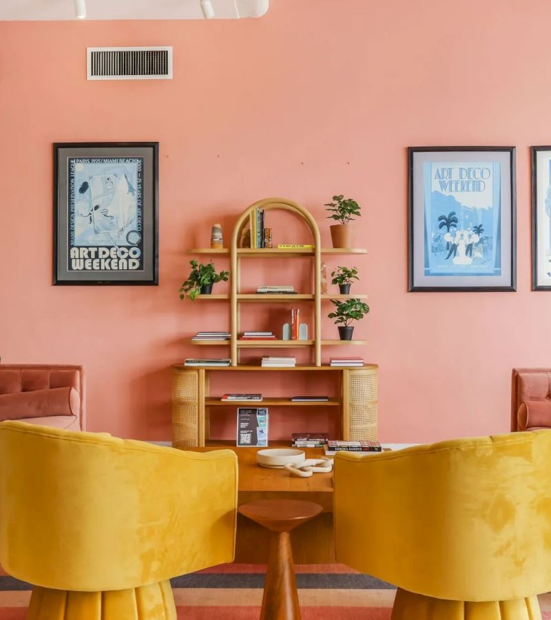A brightly colored lounge area with yellow chairs and a wooden table in front of a pink wall.