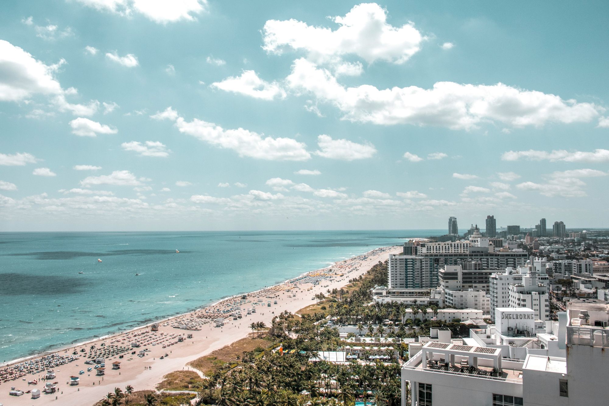 A sunny coastline with a sandy beach, turquoise ocean, and a distant city skyline under fluffy clouds.