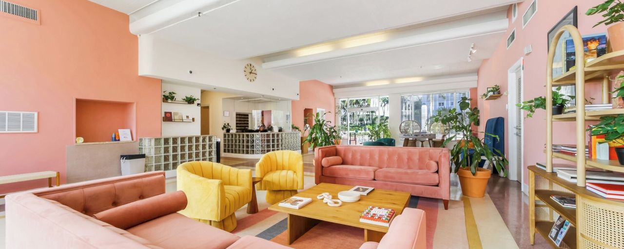 A lobby with pink walls, a pink sofa, yellow armchairs, a wooden coffee table, and a bookshelf.